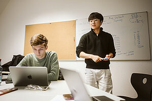 Zwei Studenten bei einer Präsentation, der linke sitzt am Laptop, der rechte steht